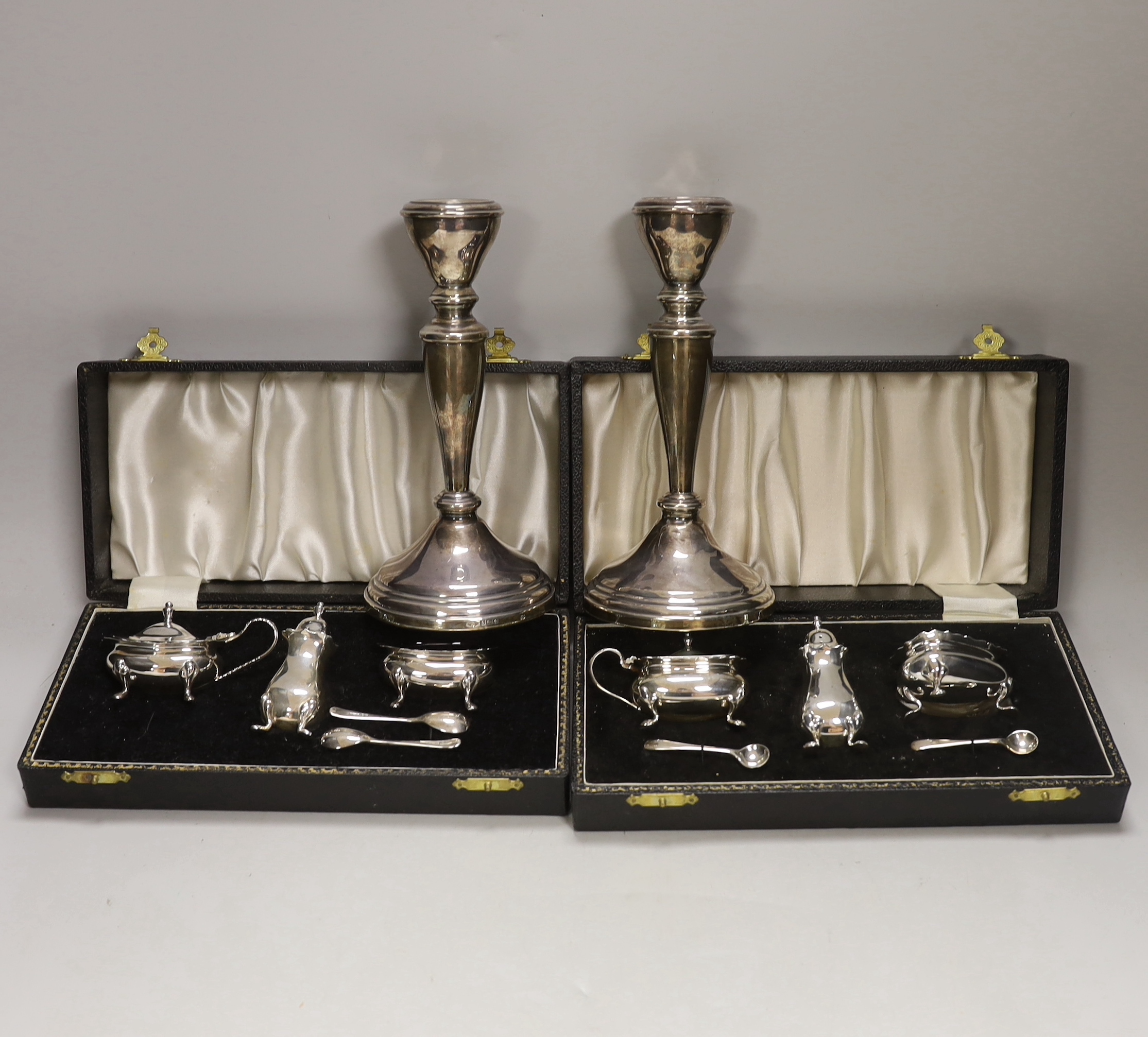 Two modern cased silver three piece cruet sets, with spoons and a pair of modern silver candlesticks, 19.5cm, weighted.